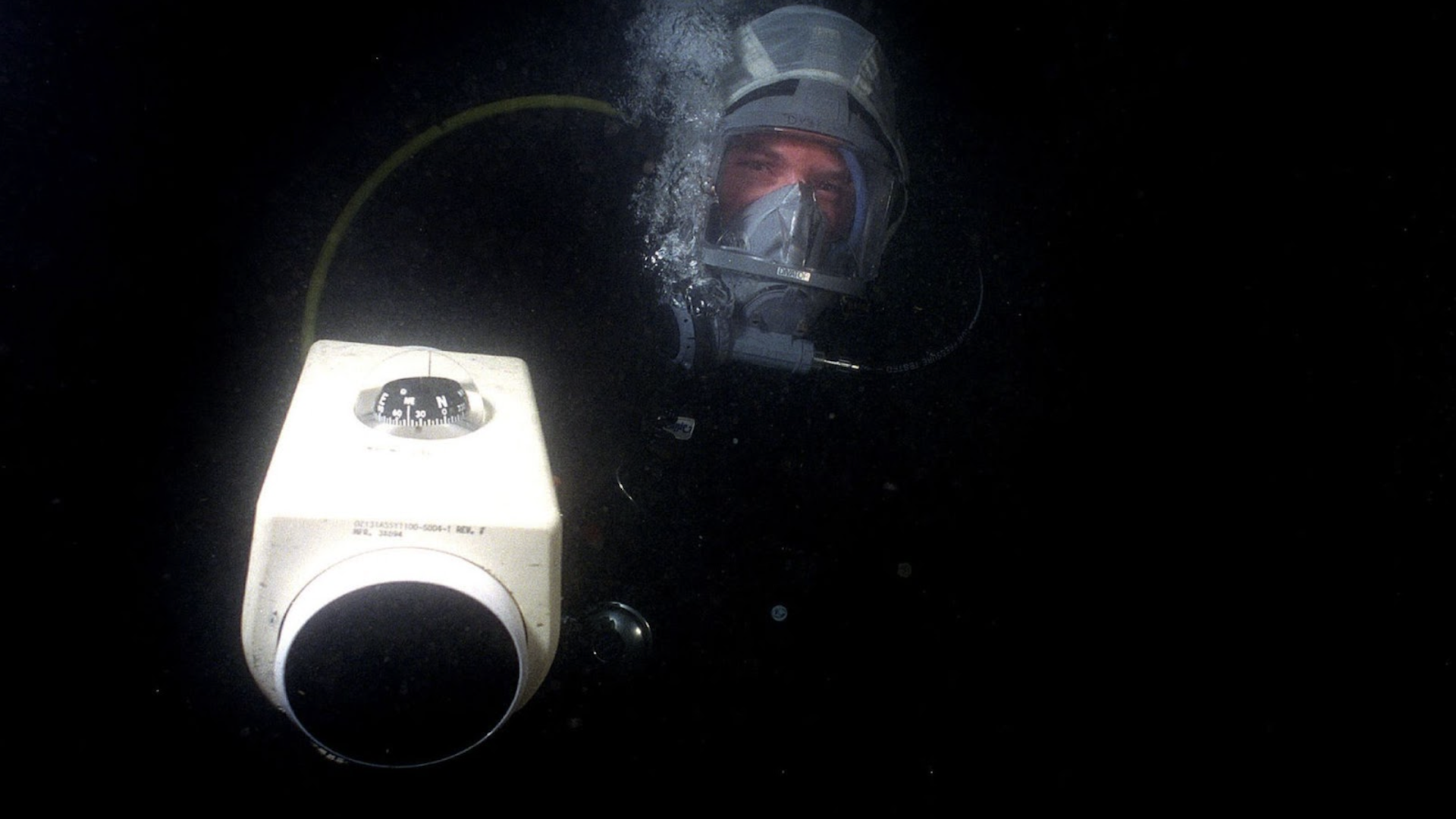 An underwater photograph of a diver wearing a full-face mask and a helmet, positioned behind a piece of navigation equipment with a visible compass